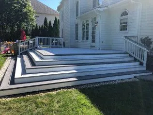White house with gray and white tiered deck and stairs, overlooking a lawn.