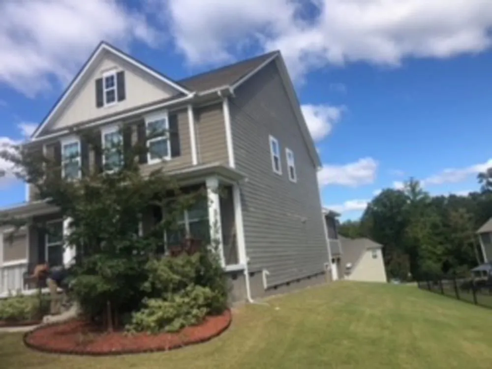 Two-story house with tan siding, black shutters, and a sloped roof under a cloudy blue sky.