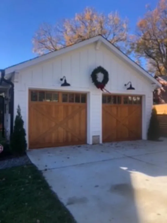 White garage with two brown wood doors, Christmas wreath, and black sconces. Concrete driveway, green grass, and trees.