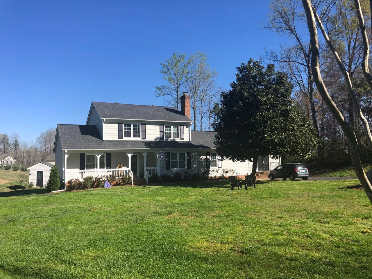 White two-story house with black shutters, chimney, and porch, on a grassy hill, under a blue sky.