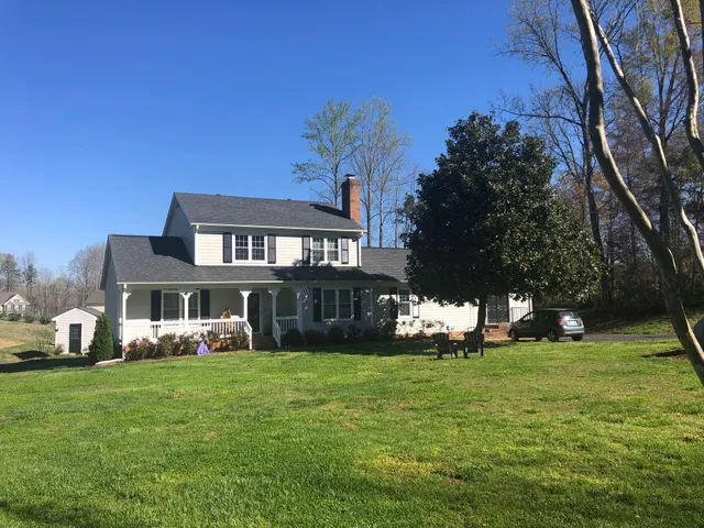 White two-story house with black shutters, chimney, and porch, on a grassy hill, under a blue sky.