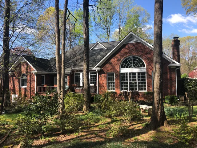 Brick house with white trim, arched window, and chimney, surrounded by trees and greenery under a blue sky.