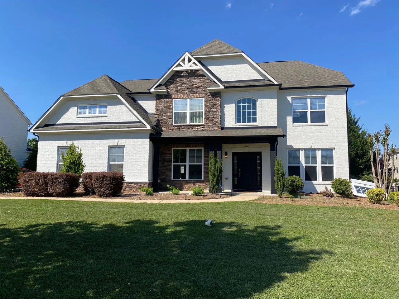 Two-story white house with brown brick accents, black door, and green lawn on a sunny day.