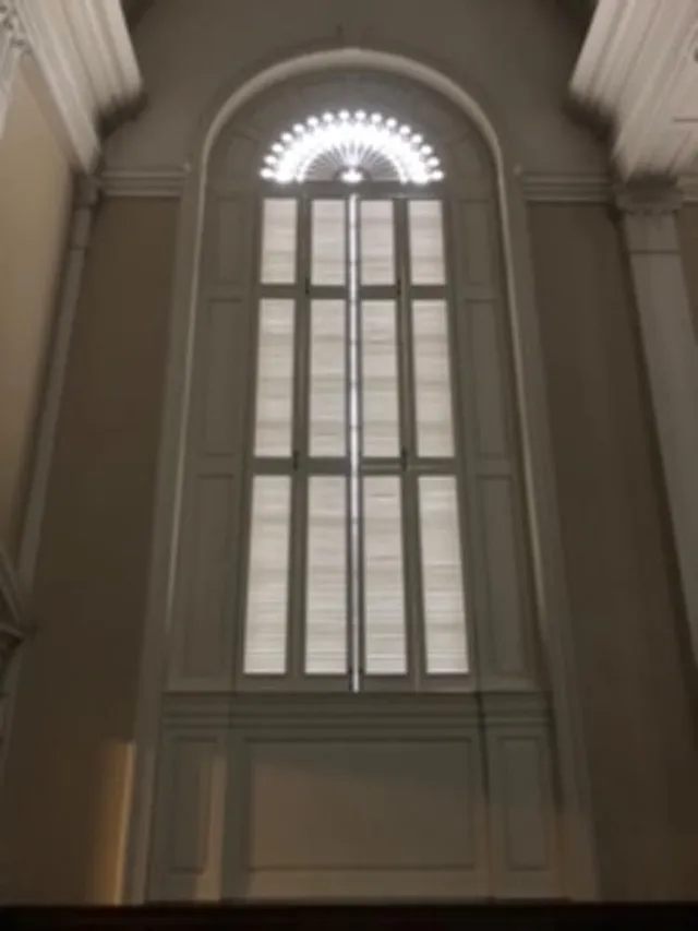 Tall arched window with white frame and blinds, in a cream-colored room.