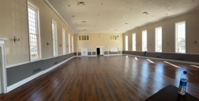 Empty, spacious hall with wood floor, tall windows, and cream-colored walls.