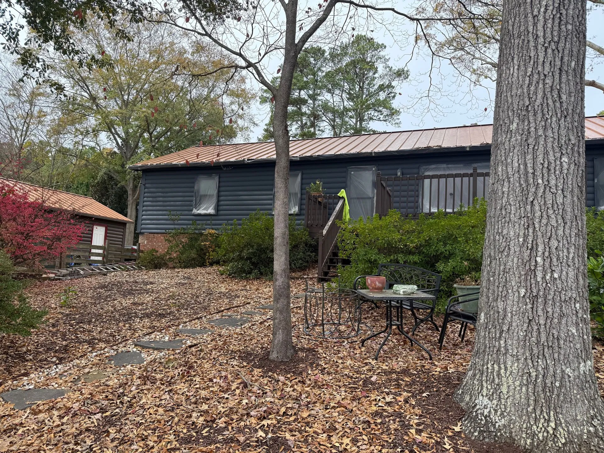 Dark green building with red roof, small side building, trees and table in yard. Autumn leaves cover ground.