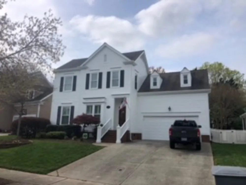 White two-story house with black shutters, dormers, and a garage. Black truck parked in driveway. Green lawn and trees.