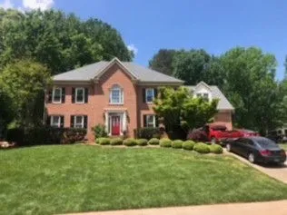 Two-story brick house with black shutters, red door, green lawn, trees, and parked cars.