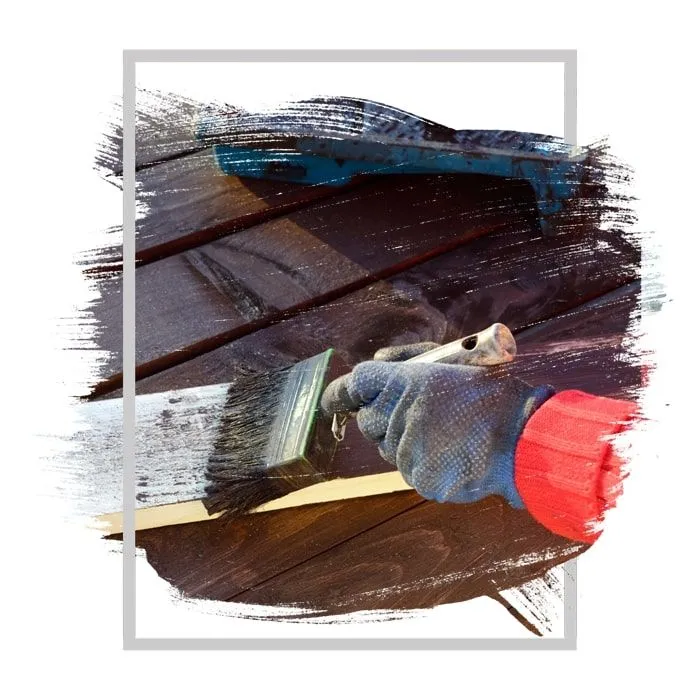Person with gloved hand painting wooden planks with a brush; dark stain applied.
