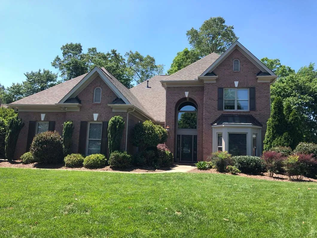 Brick house with a brown roof, green lawn, and shrubbery under a blue sky.