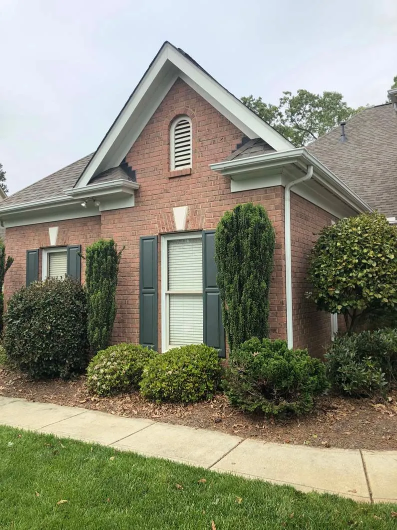 Brick house with shutters and landscaping, under a cloudy sky.