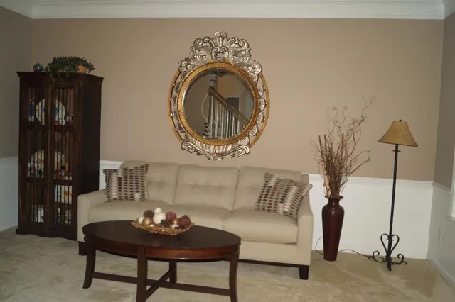 Living room with tan walls, white wainscoting, beige sofa, round mirror, dark wood cabinet, coffee table, lamp, and vase.