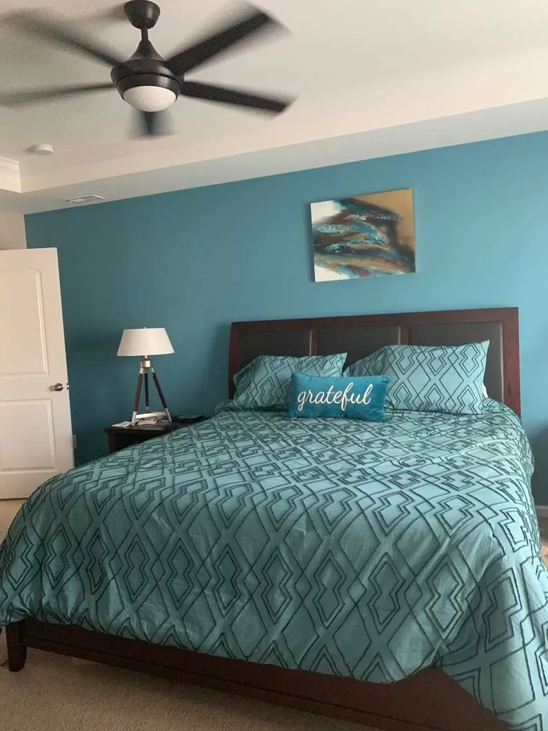 Bedroom with teal accent wall, bed, artwork, lamp, and ceiling fan.