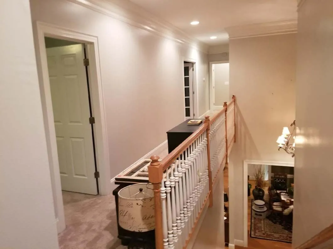 Hallway with a staircase, doors, and a decorative railing. Beige walls, wood accents, and overhead lighting.