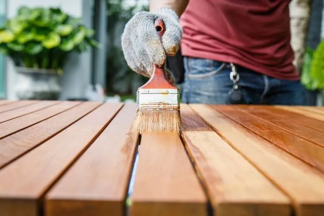 Person applying varnish to a wooden table with a paintbrush outdoors.