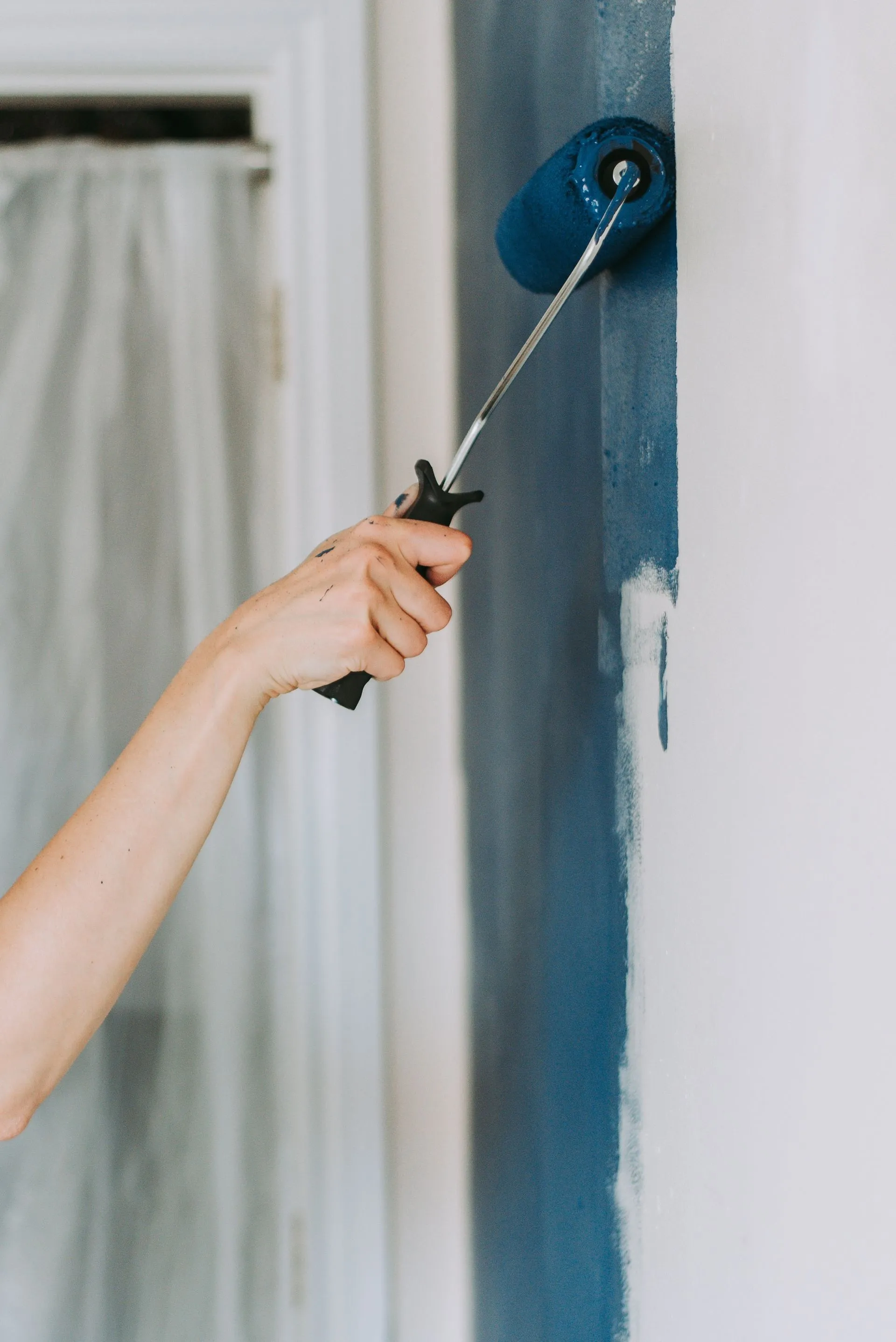 A person paints a wall blue with a roller; interior setting, white wall, window.