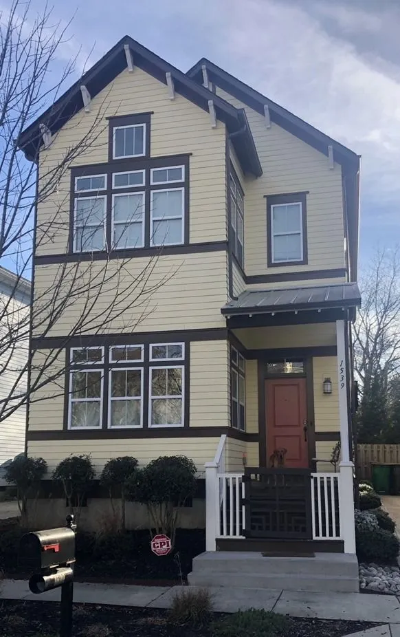 Two-story yellow house with brown trim and red door, front porch, bushes, and mailbox.